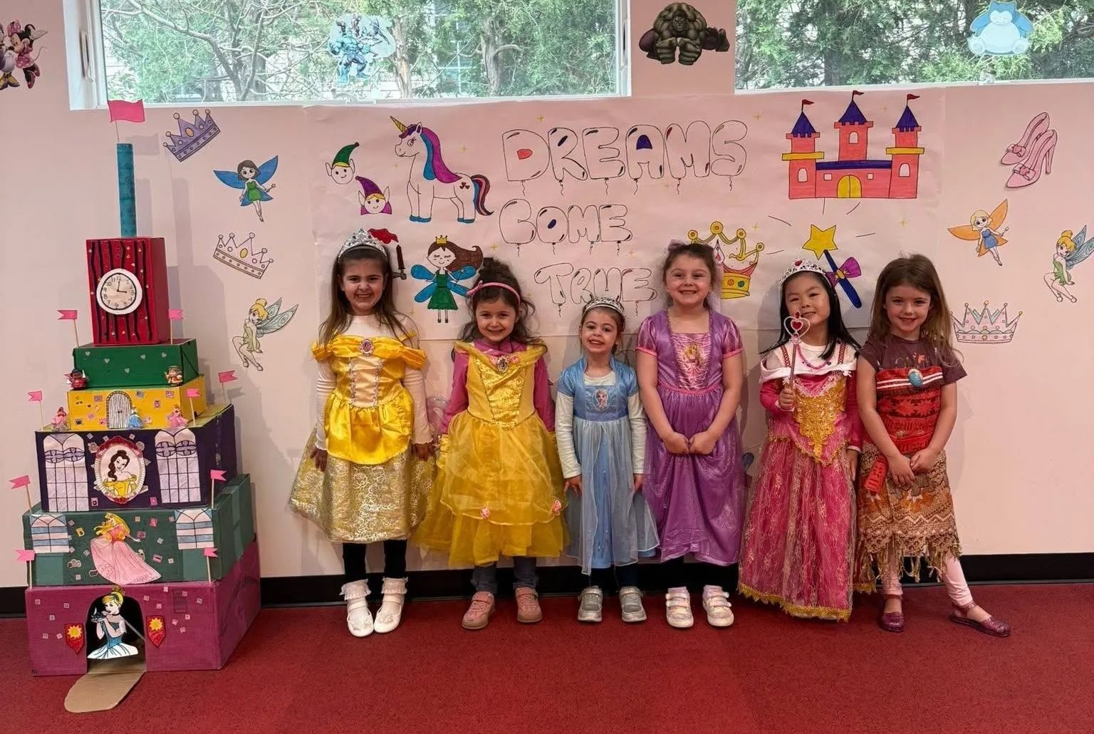 Children in princess costumes stand in front of a decorated wall; a cardboard castle is in front.