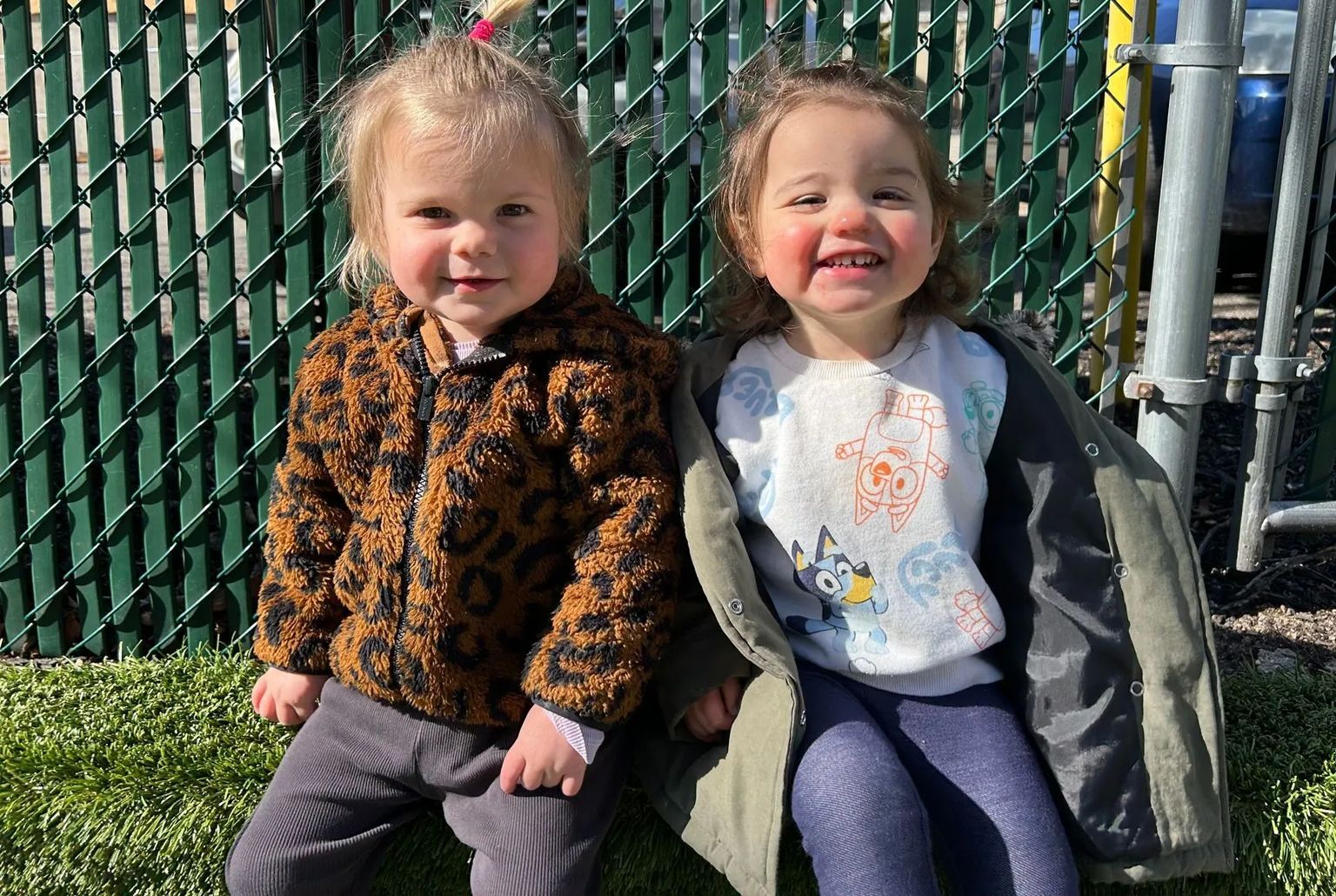 Two young children smiling and sitting next to each other outside. One in leopard jacket, the other in a light green coat.