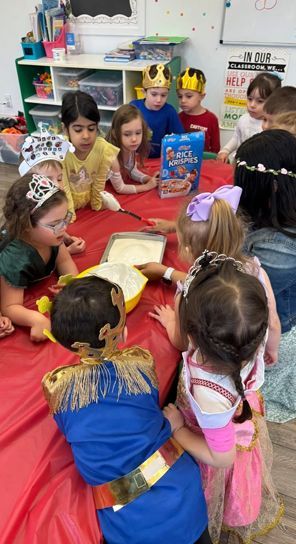 Children in costumes gather around a table, possibly baking or crafting, in a classroom.