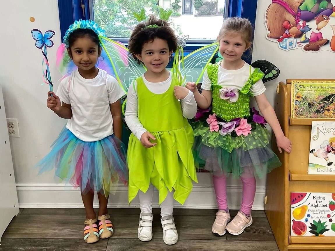 Three young children dressed as fairies posing by a window; colorful tutus, wings, wands, happy expressions.