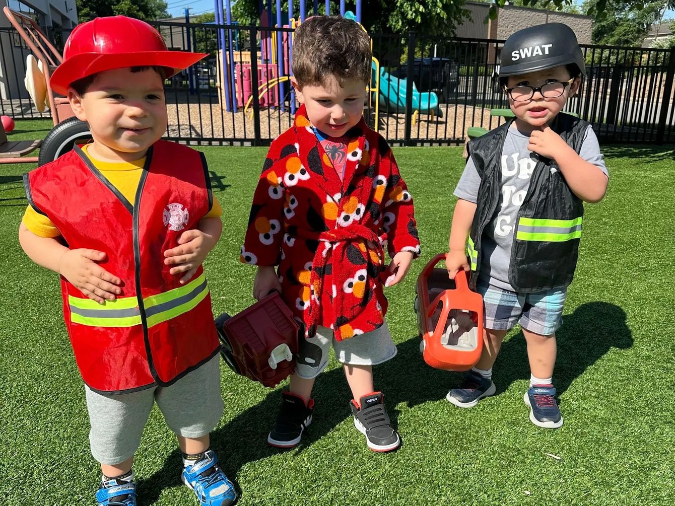 Three young children in costumes, including a firefighter, in a grassy area.