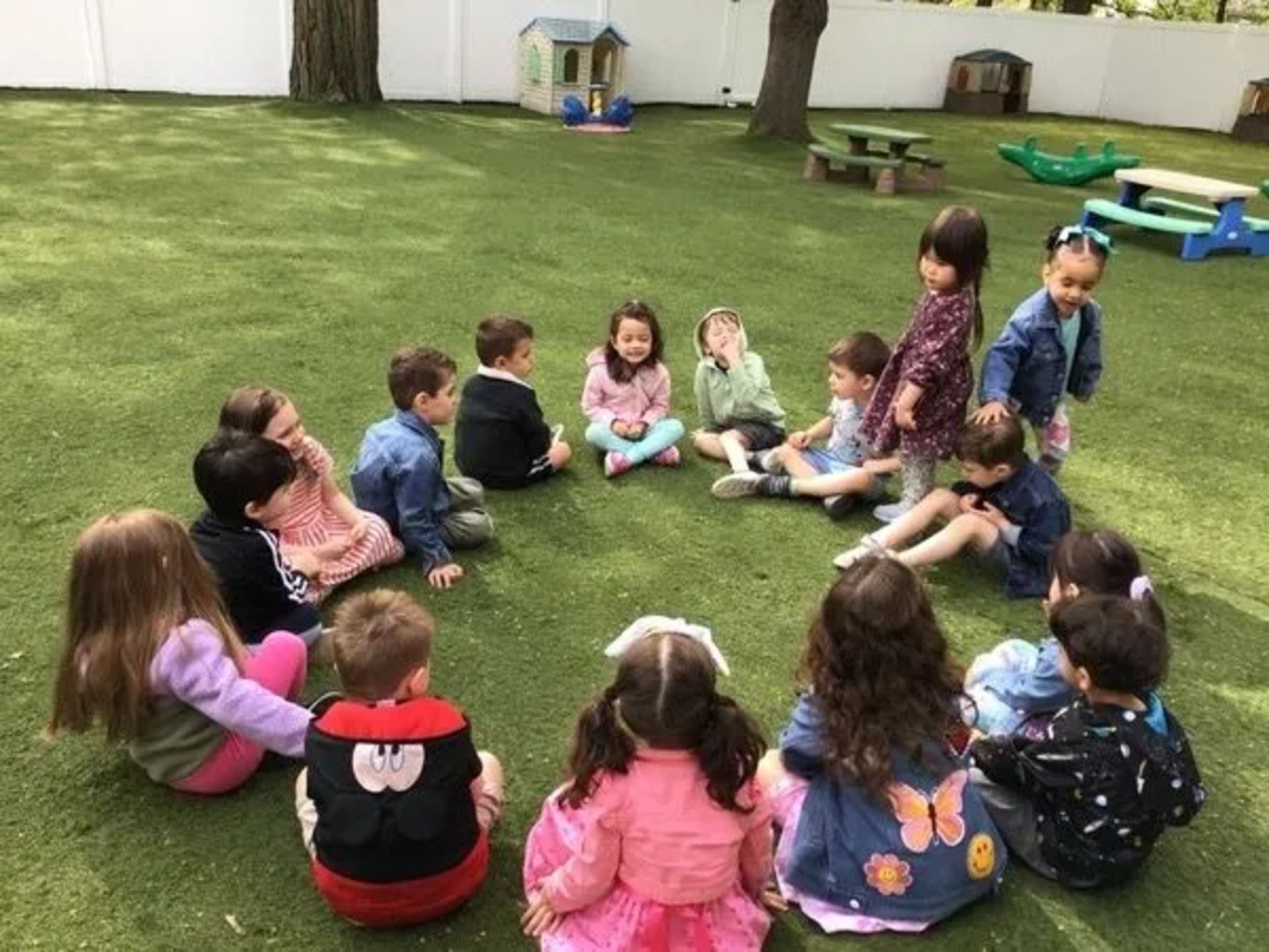 Children sitting in a circle on green grass, outside. Some are talking and looking at each other.