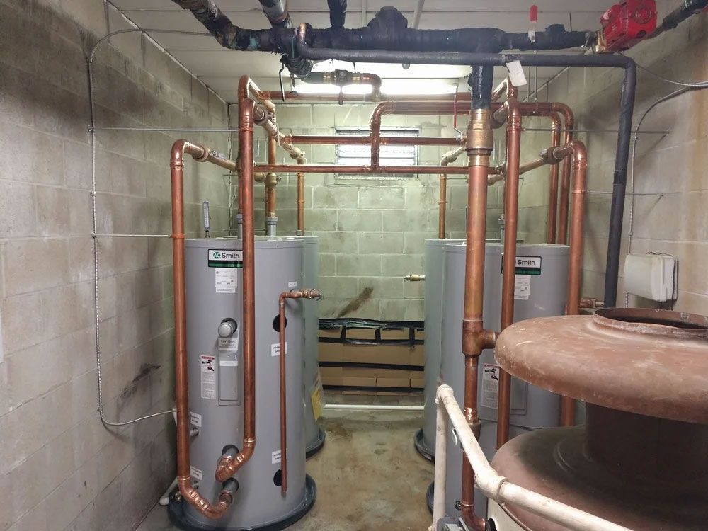 Water heater room with copper pipes and gray tanks against a block wall.