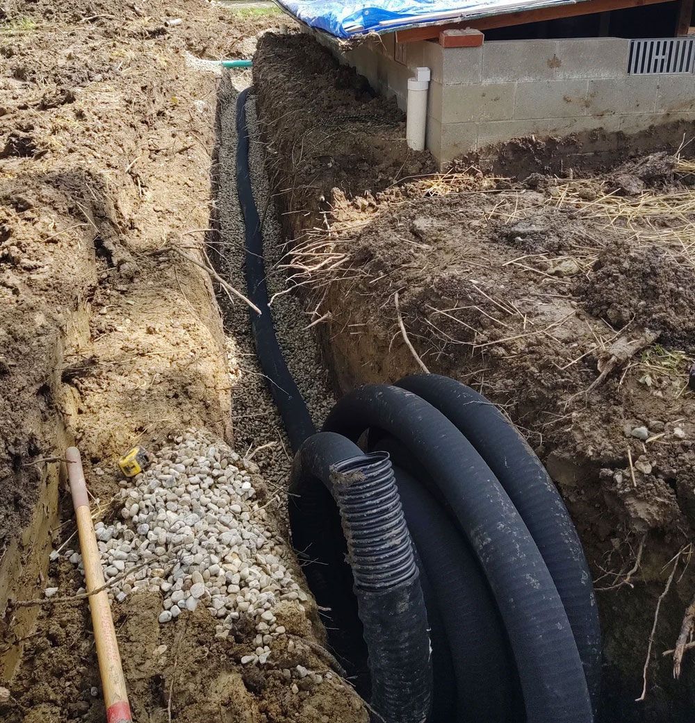 Trench with black drainage pipe being installed next to a building foundation; soil and gravel visible.