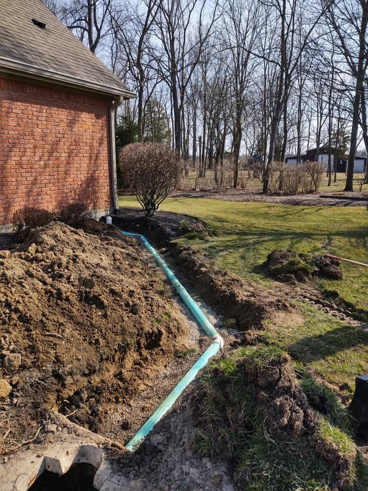 Trench dug along a brick house, containing green pipe. Dirt mound, grassy yard, bare trees.
