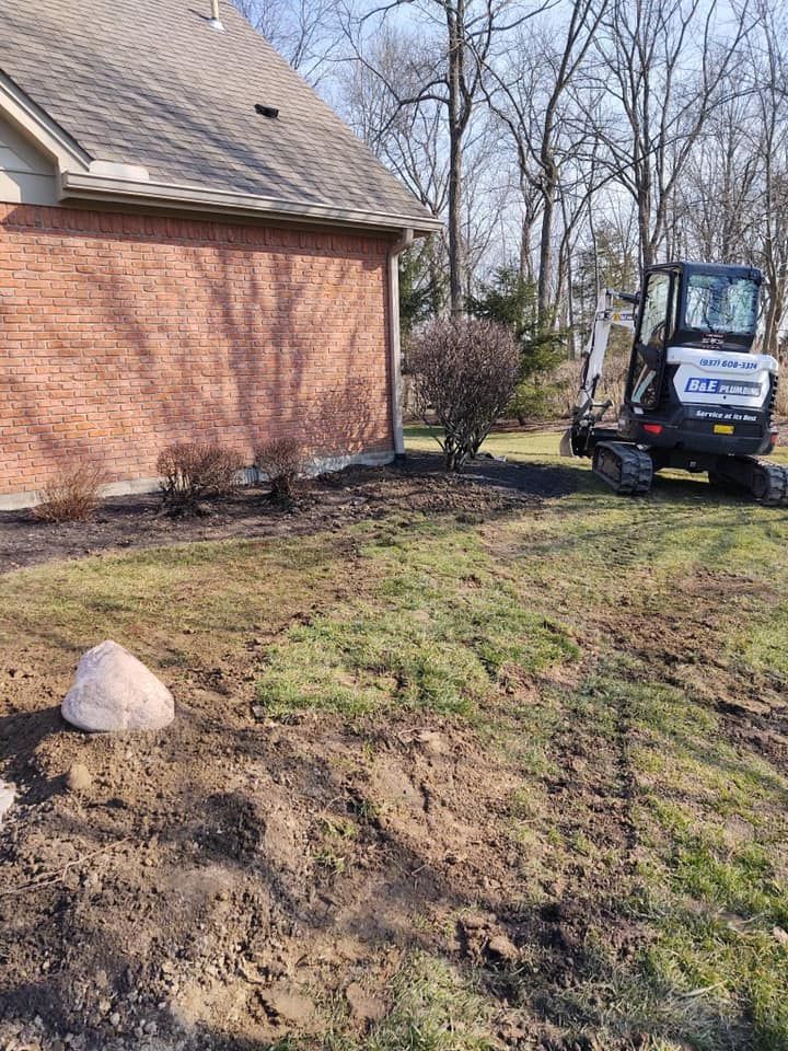A Bobcat excavator working on landscaping near a brick house and a small bush in a yard.
