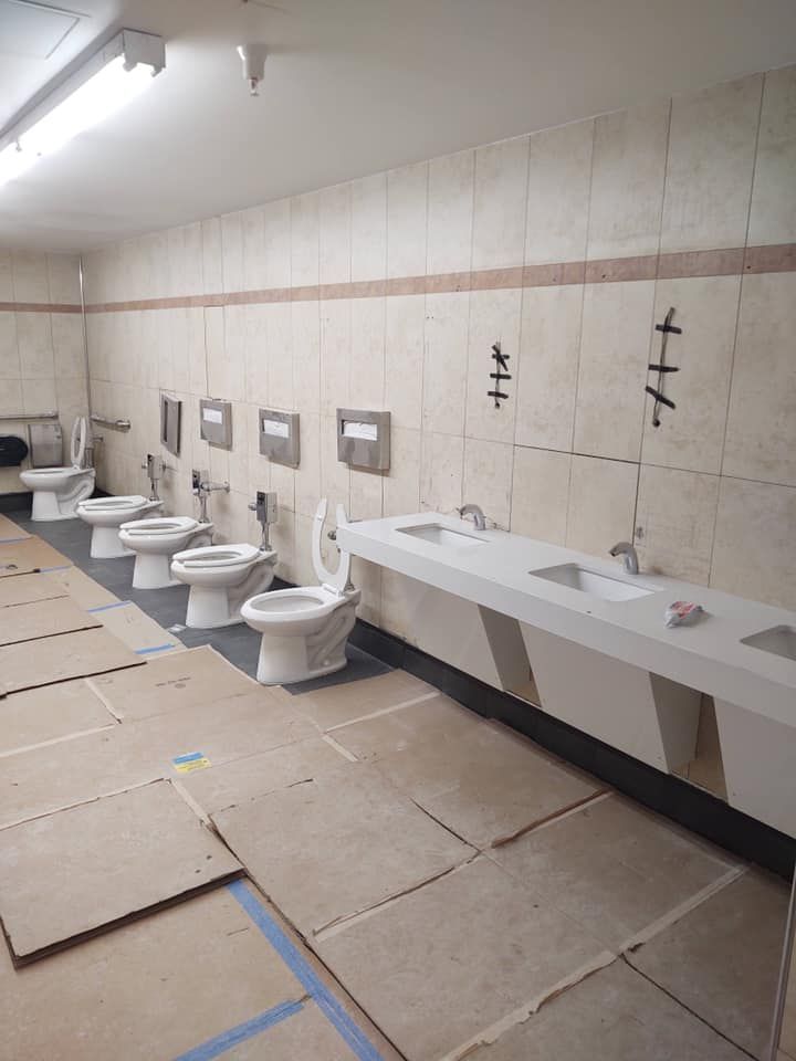 Bathroom with multiple white toilets, sinks, beige tile walls, and flooring covered in cardboard.