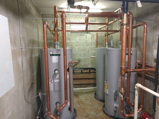 Three silver water heaters with copper piping in a concrete-walled utility room.