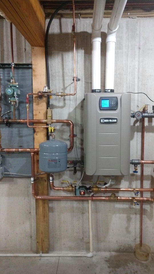 A wall-mounted gray tankless water heater with copper pipes in a basement setting.