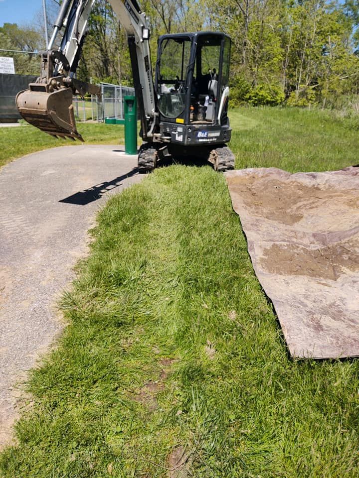 Mini excavator on a grassy strip next to a walking path; operator's view.