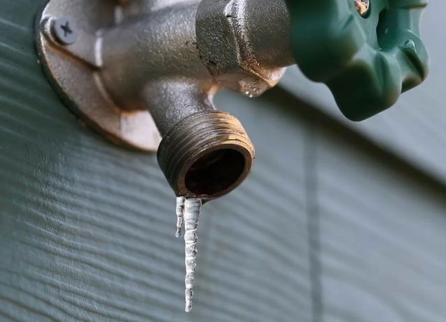 An outdoor faucet with an icicle forming on its spout, mounted on a green wall.