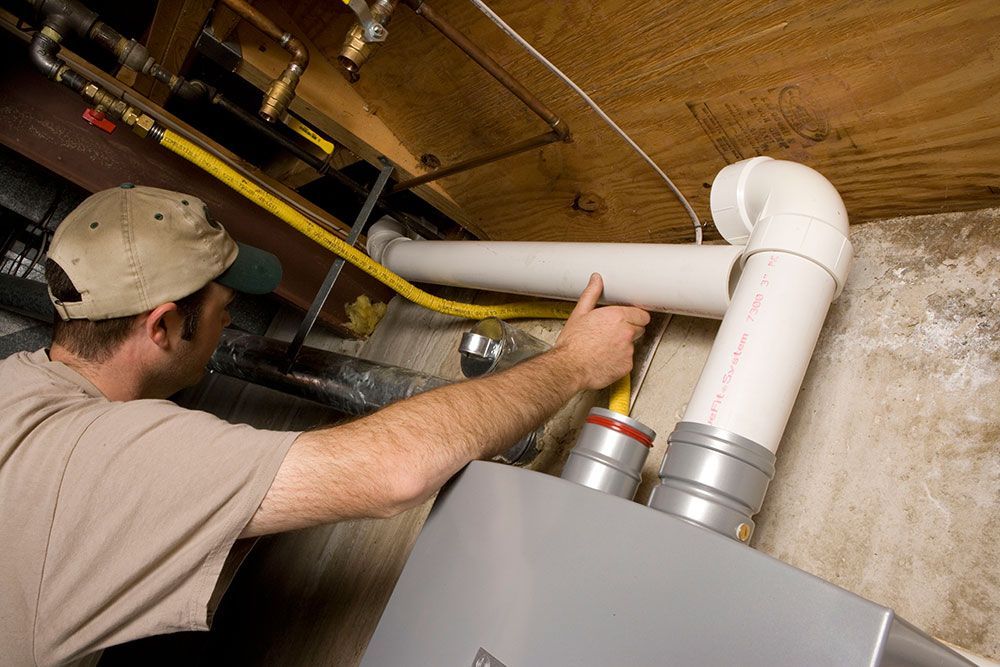 Man installing pipes to a furnace in a basement, with yellow gas line visible.