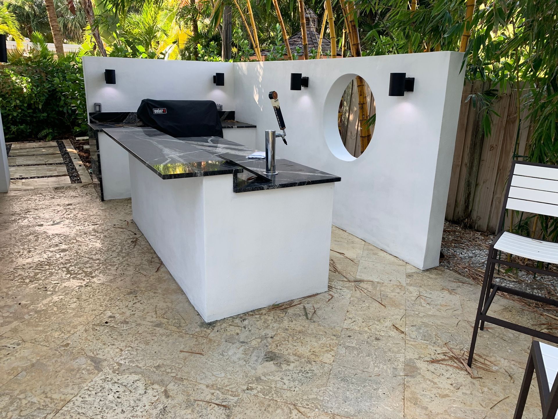 Outdoor white barbecue kitchen with black countertops and sink on a patio, surrounded by trees