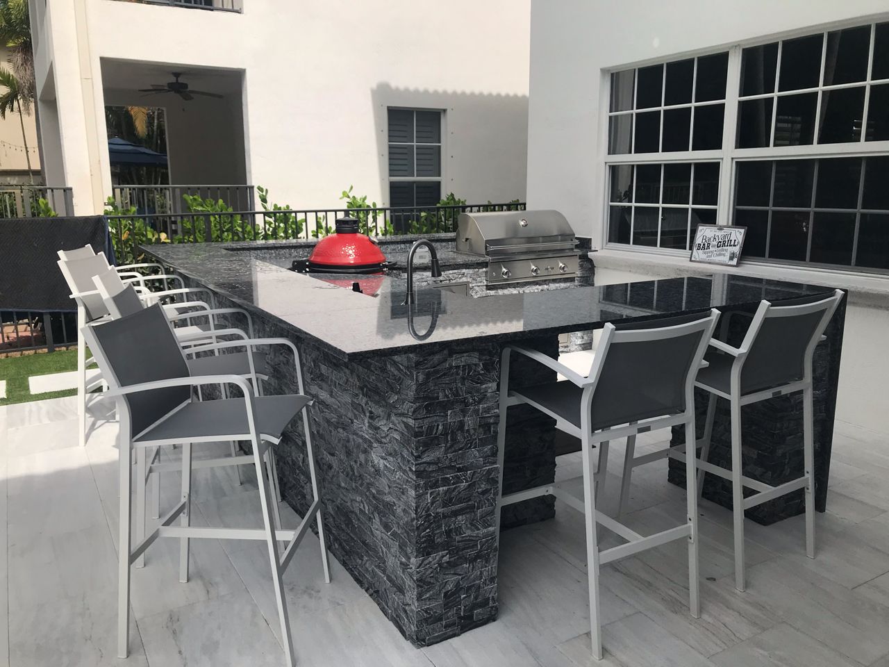 Outdoor patio dining table with gray chairs, a red grill, and a black stone base beside a building