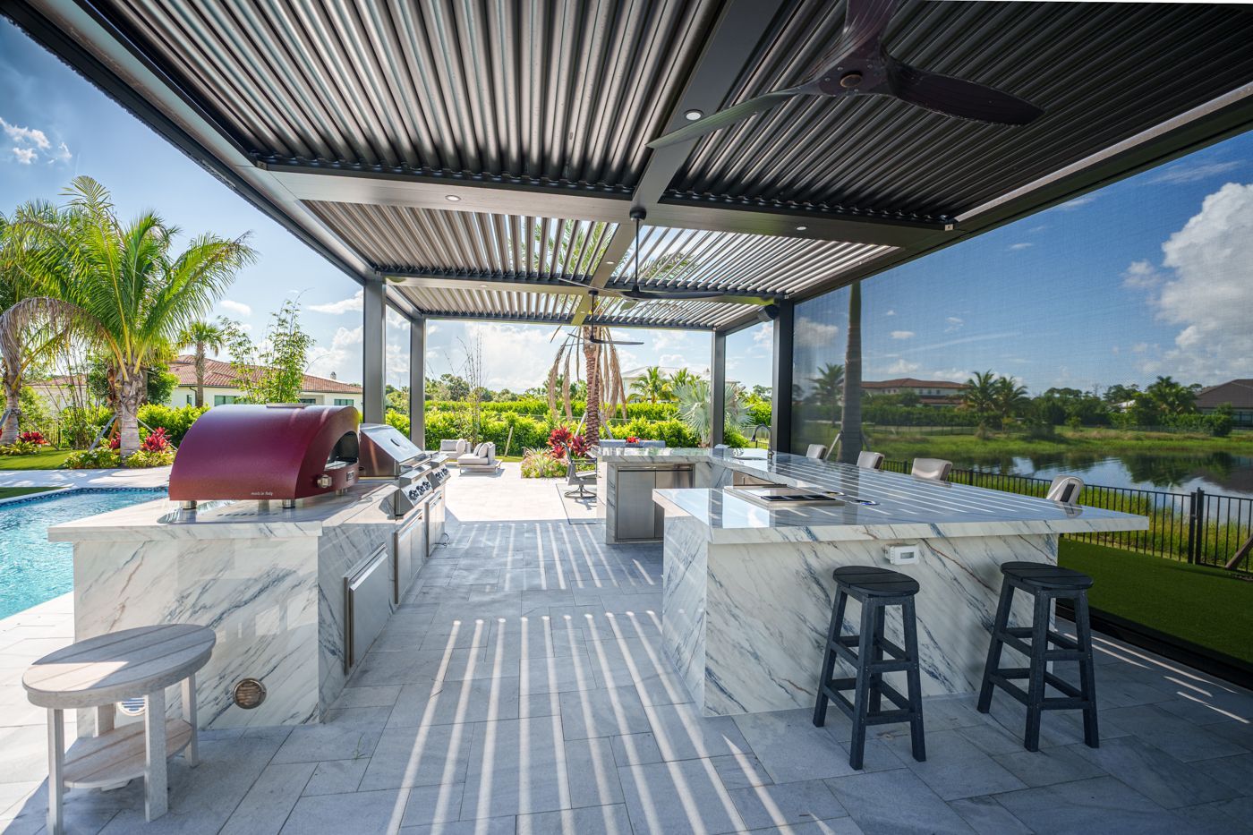 Modern outdoor patio with dining table, grill, bar stools, and poolside lake view under a slatted pergola
