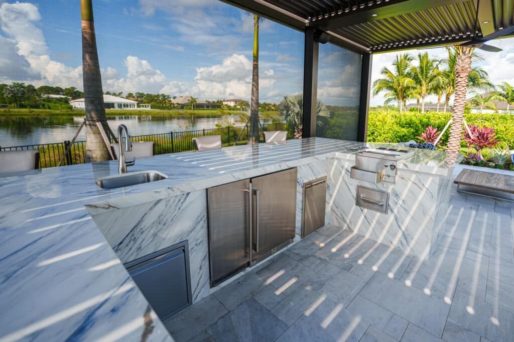 Modern outdoor kitchen with marble countertops, bar seating, and a lake view under a shaded pergola