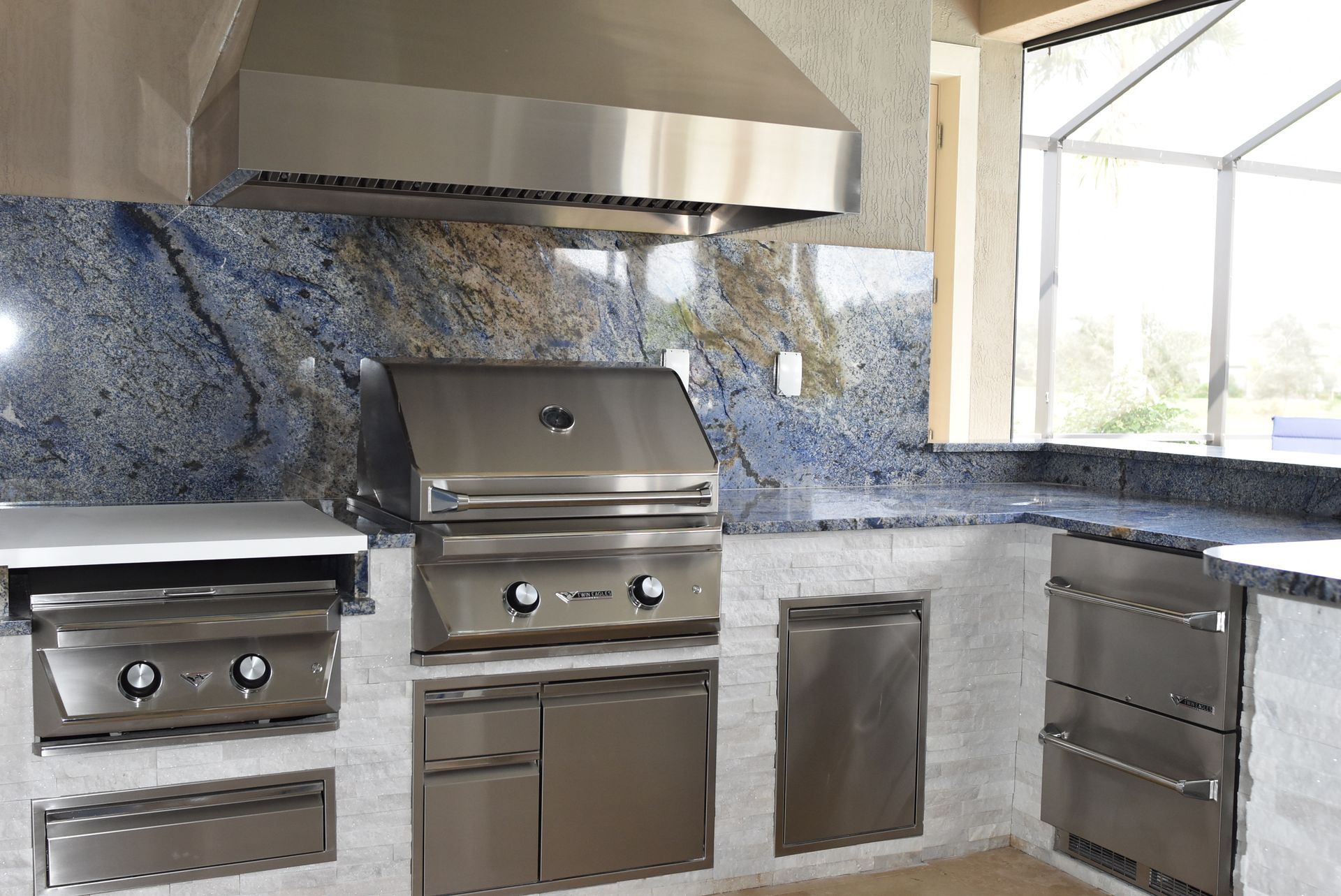 Modern outdoor kitchen with stainless steel grills, cabinets, and blue stone backsplash under a range hood.