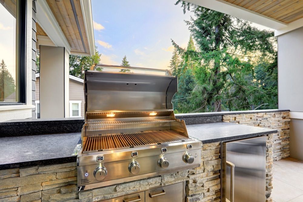 Outdoor stainless steel grill on a covered patio with stone counters and trees beyond