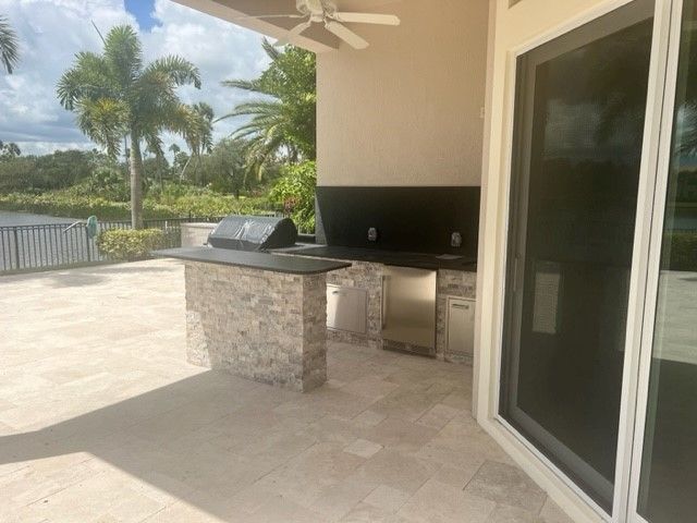Covered patio with outdoor kitchen, stone bar, ceiling fan, sliding glass door, and palm trees beyond