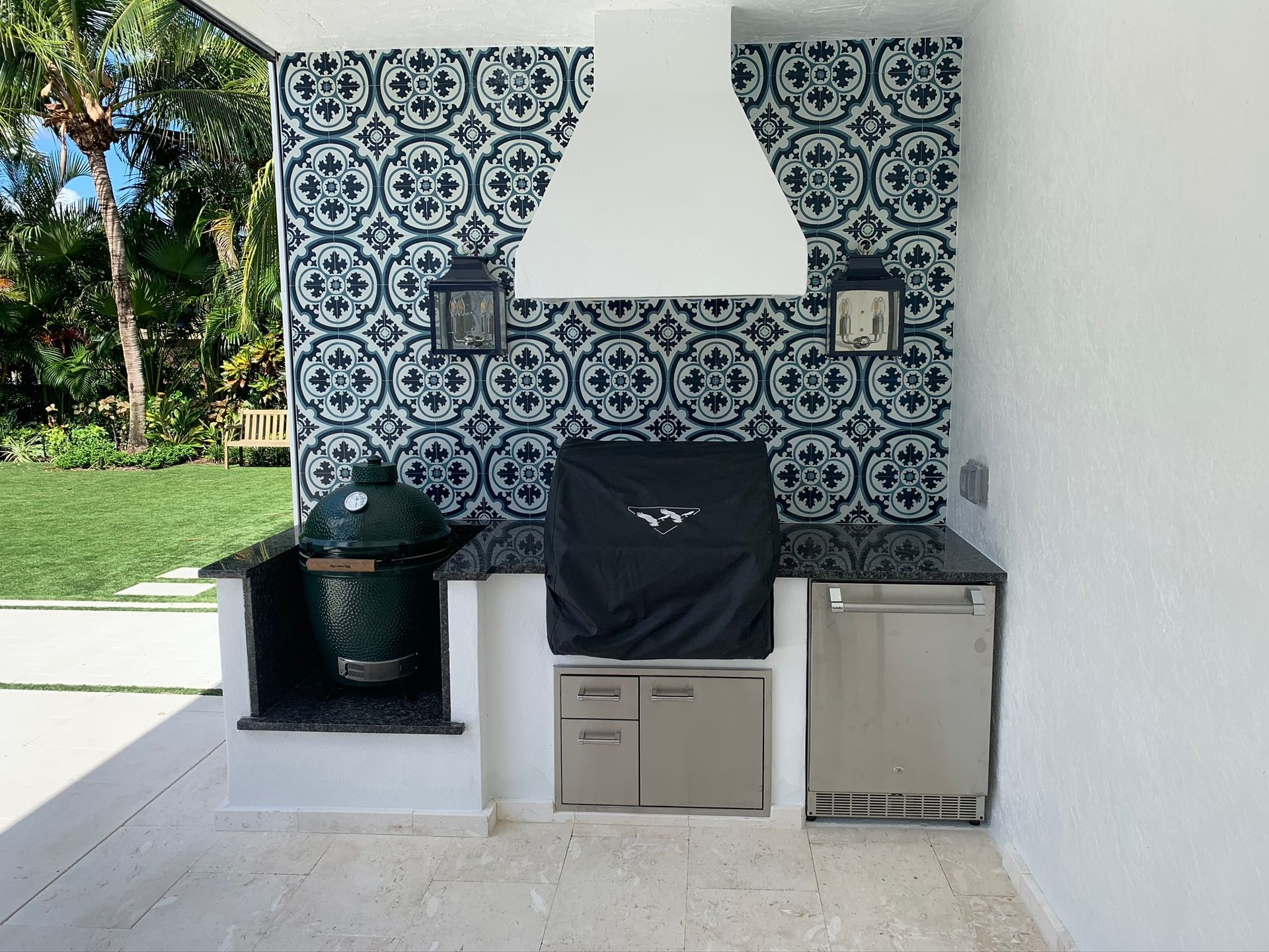 Outdoor patio grill area with patterned tile backsplash, white hood, black grill, cabinets, and sink