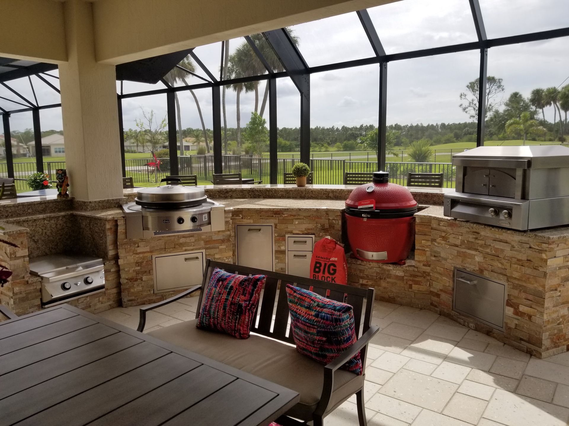 Covered outdoor patio with built-in grill, red smoker, and cushioned bench overlooking a green lawn.