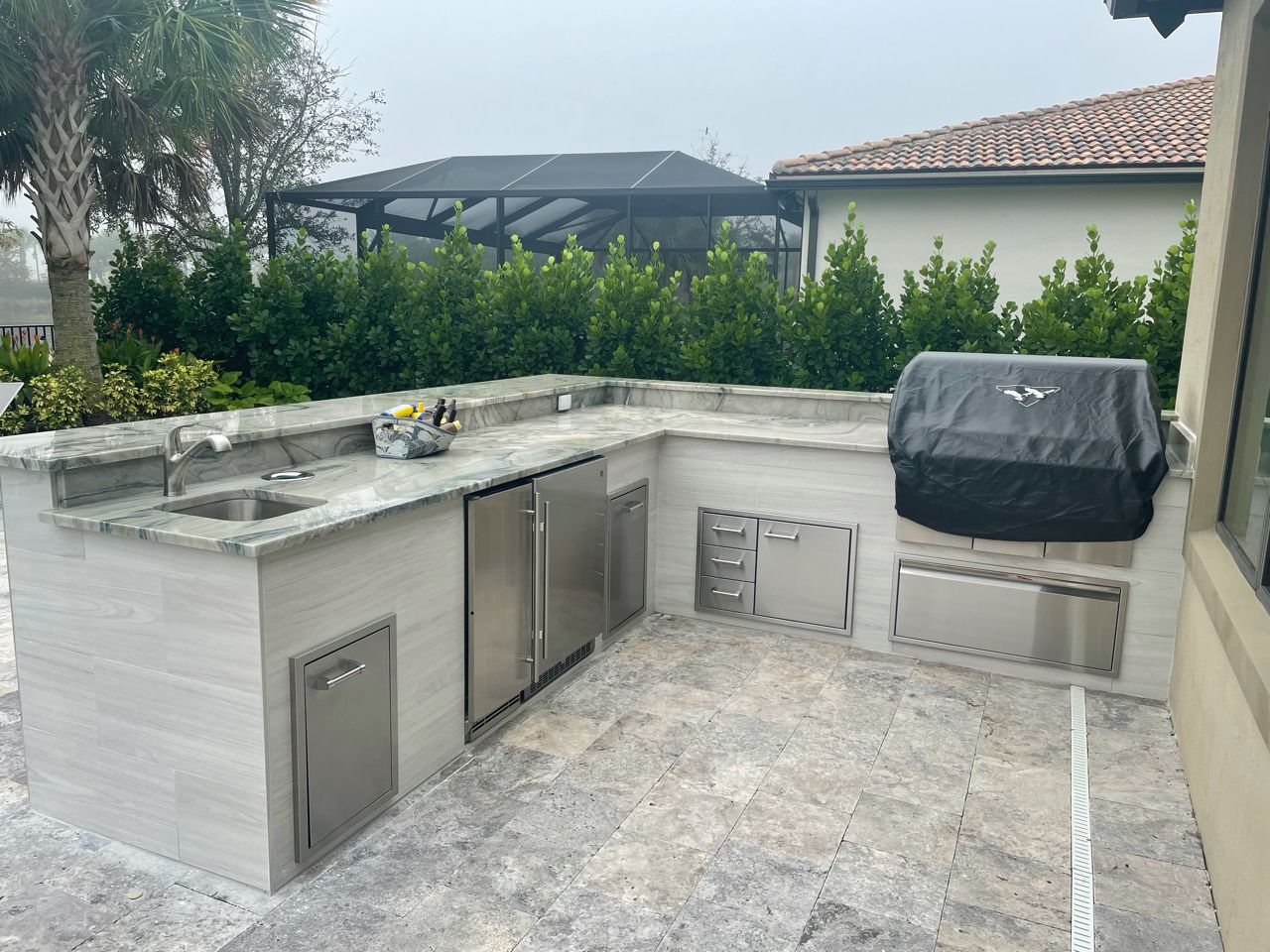 Outdoor patio kitchen with stone counters, stainless cabinets, sink, and covered grill in a backyard