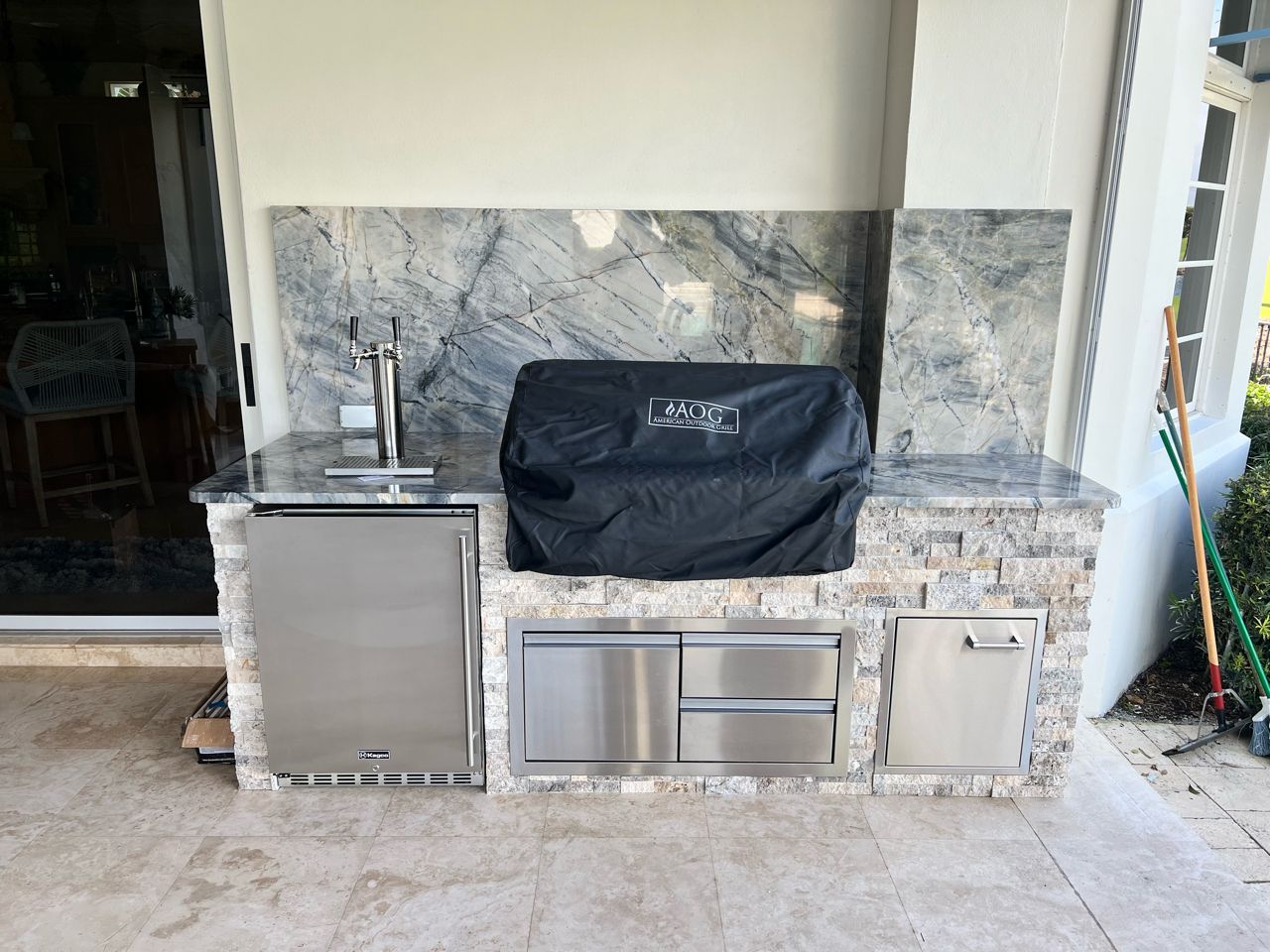 Outdoor built-in stainless steel grill and counter against a stone wall, covered with a black grill cover.
