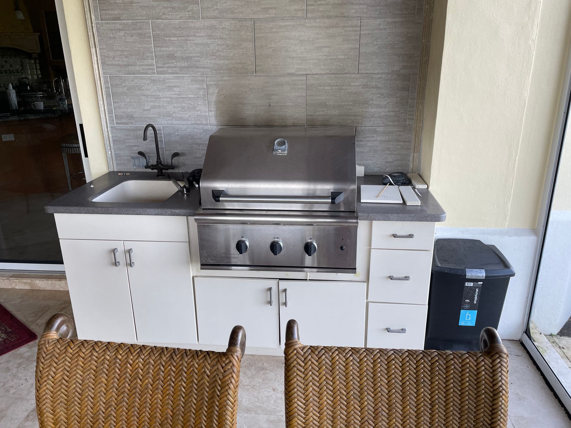 Outdoor grill station with stainless steel grill, white cabinets, sink, and wicker chairs in front
