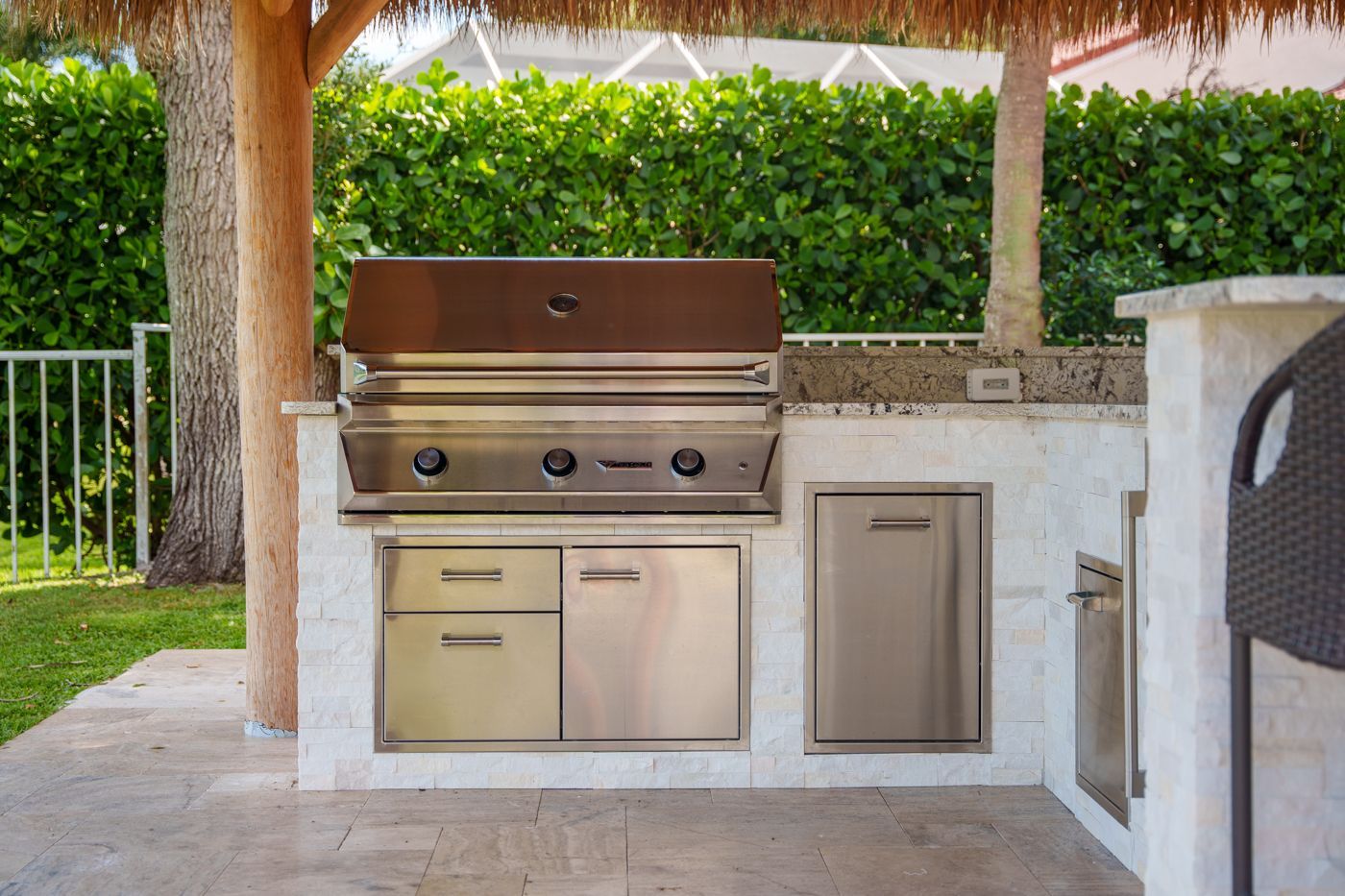 Outdoor stainless steel grill and storage cabinets under a covered patio with stone countertop and green hedge backdrop