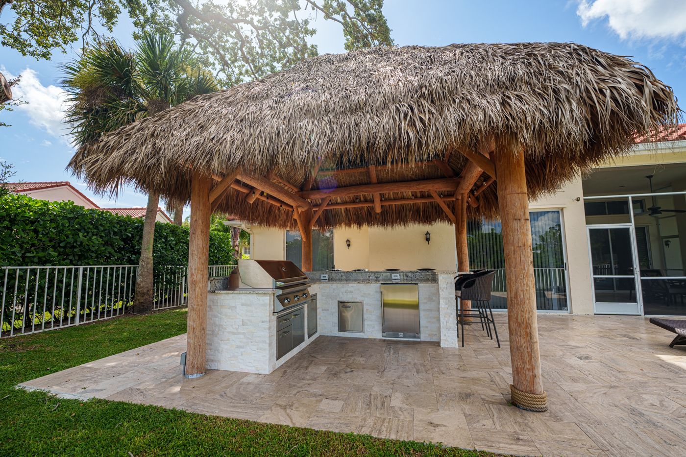 Thatched outdoor patio with a built-in grill and bar stools beside a lawn and poolside glass doors