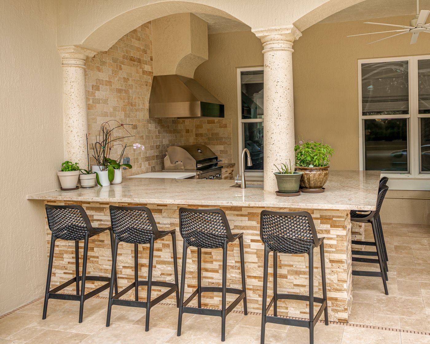 Outdoor stone bar with four black stools in a beige patio kitchen, framed by arched columns.