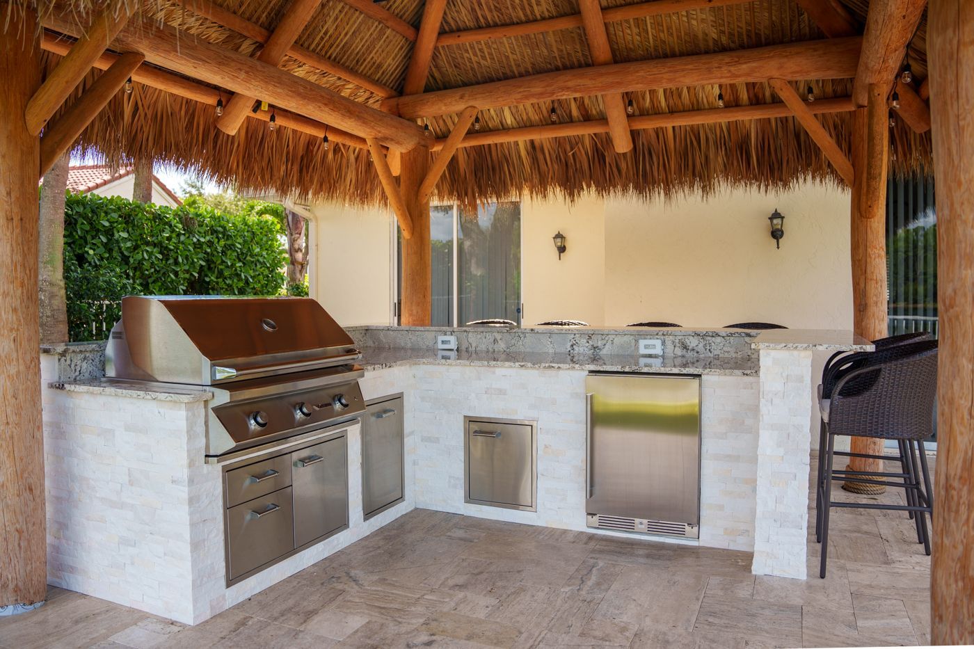 Outdoor thatched-roof kitchen with stainless grill, sink, and stone countertop under a pergola