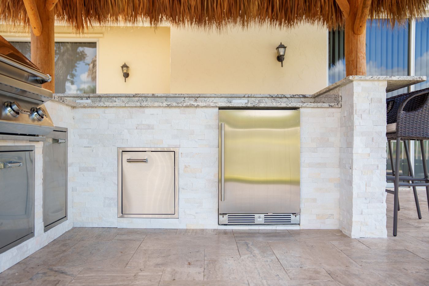 Outdoor stone kitchen with stainless appliances, built-in grill, and thatched roof bar counter