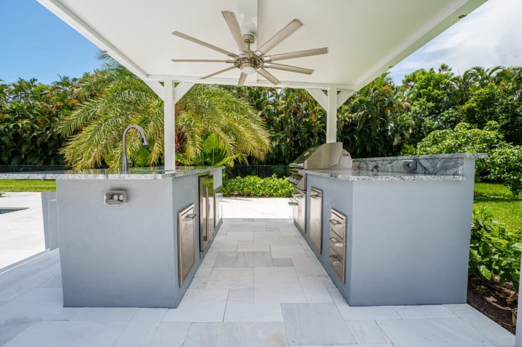 Outdoor kitchen with stainless steel grills under a white pergola, surrounded by tropical greenery