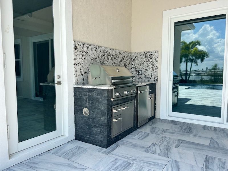 Outdoor patio with built-in grill, white marble tile floor, and glass doors overlooking a pool area