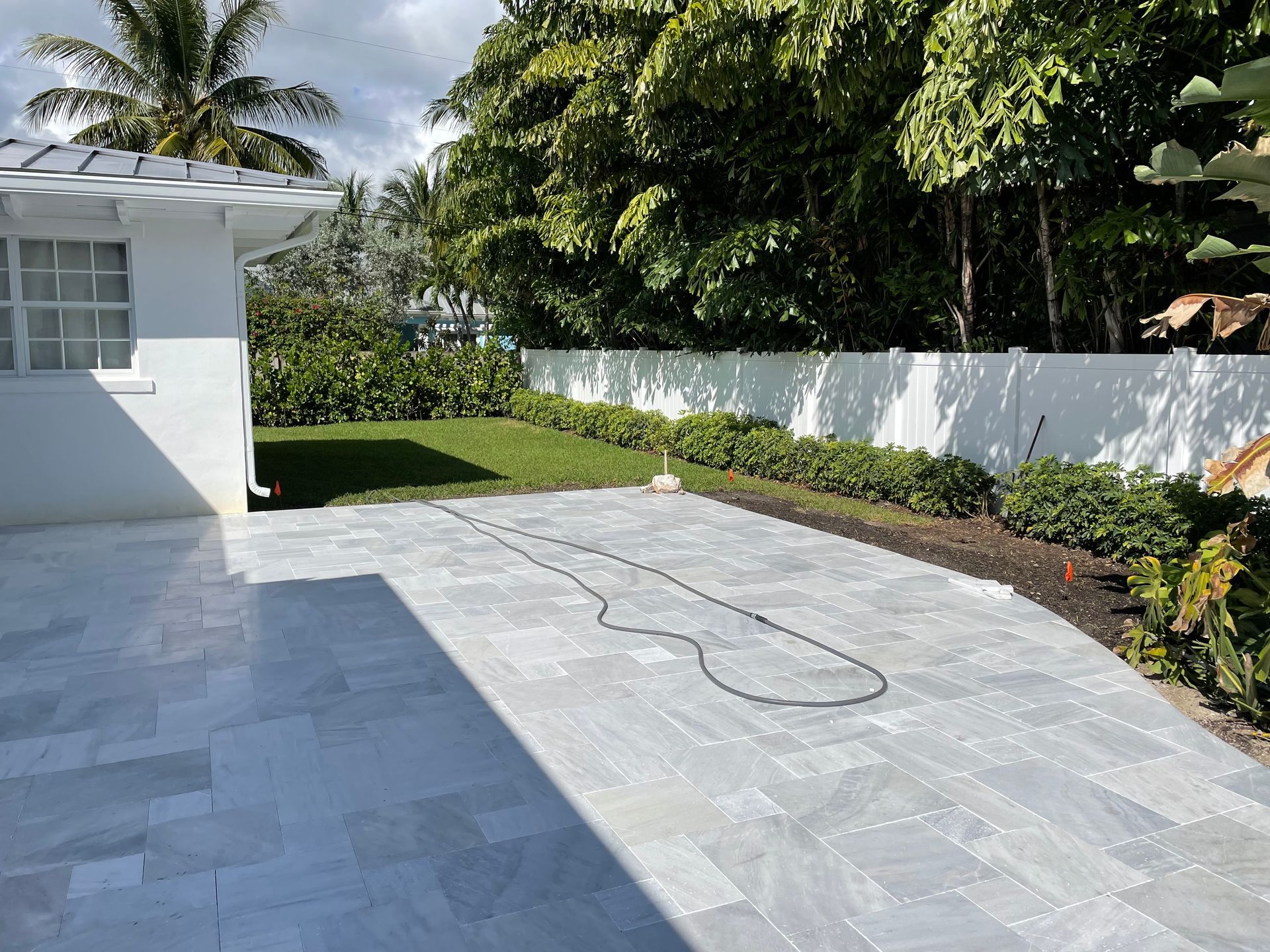 Concrete patio beside a white house, bordered by grass, hedges, and a white fence under bright sunlight