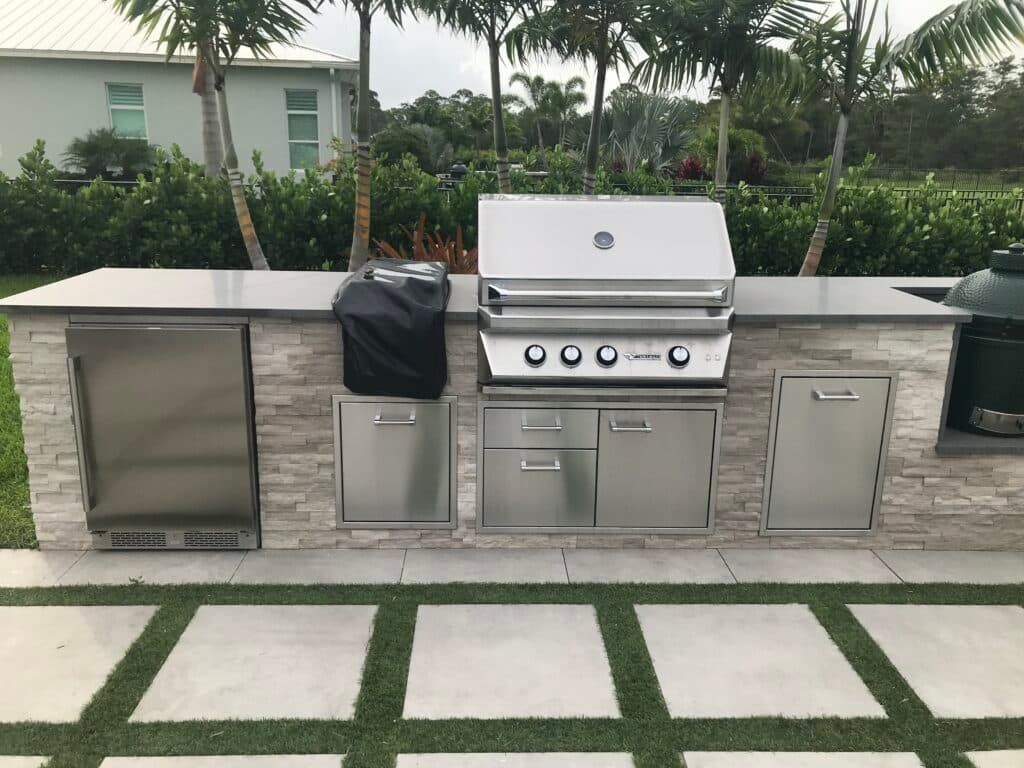Outdoor stainless steel grill island with cabinets and a covered side appliance in a landscaped patio.