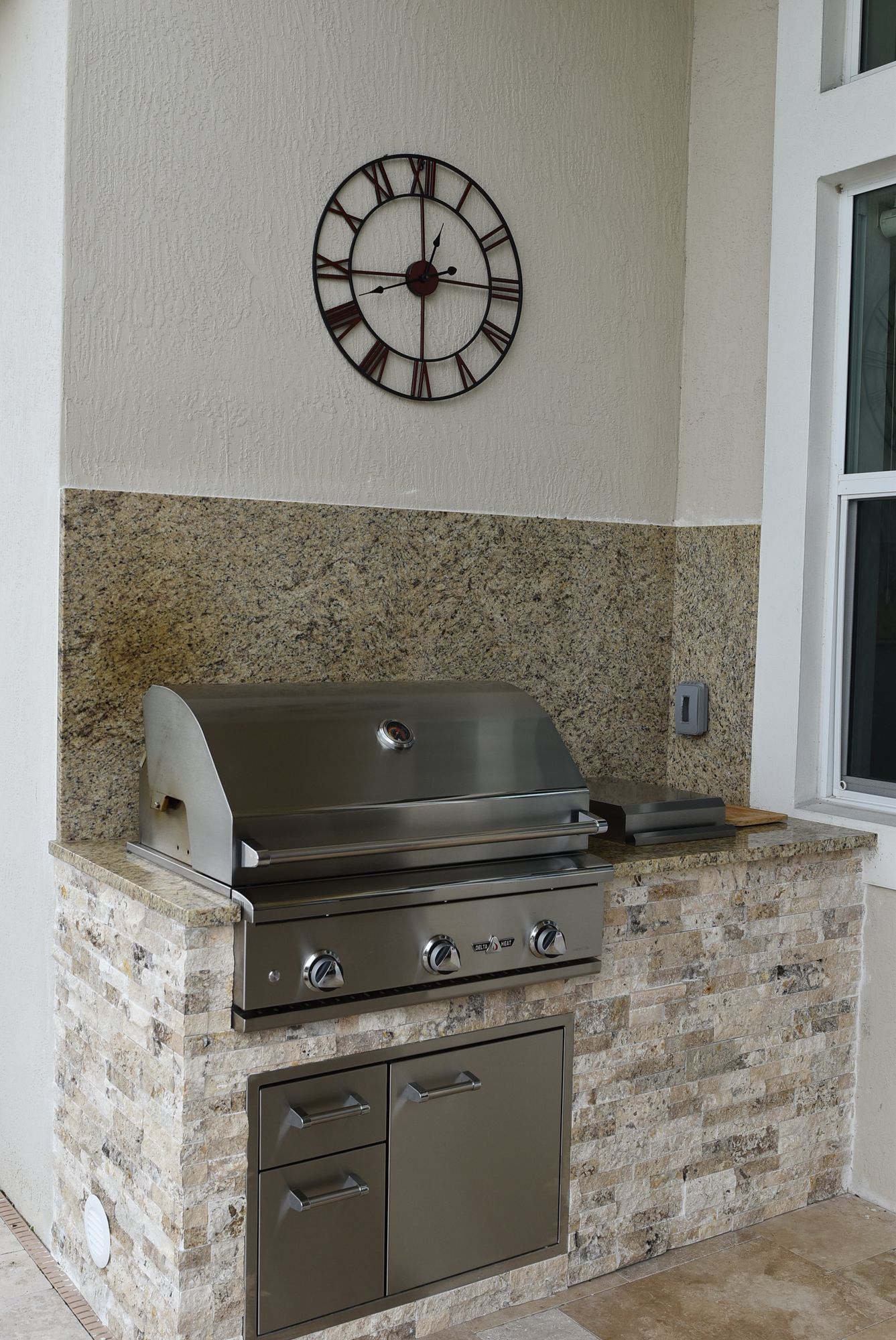Outdoor stone grill with stainless steel lid beneath a wall clock beside a window on a patio