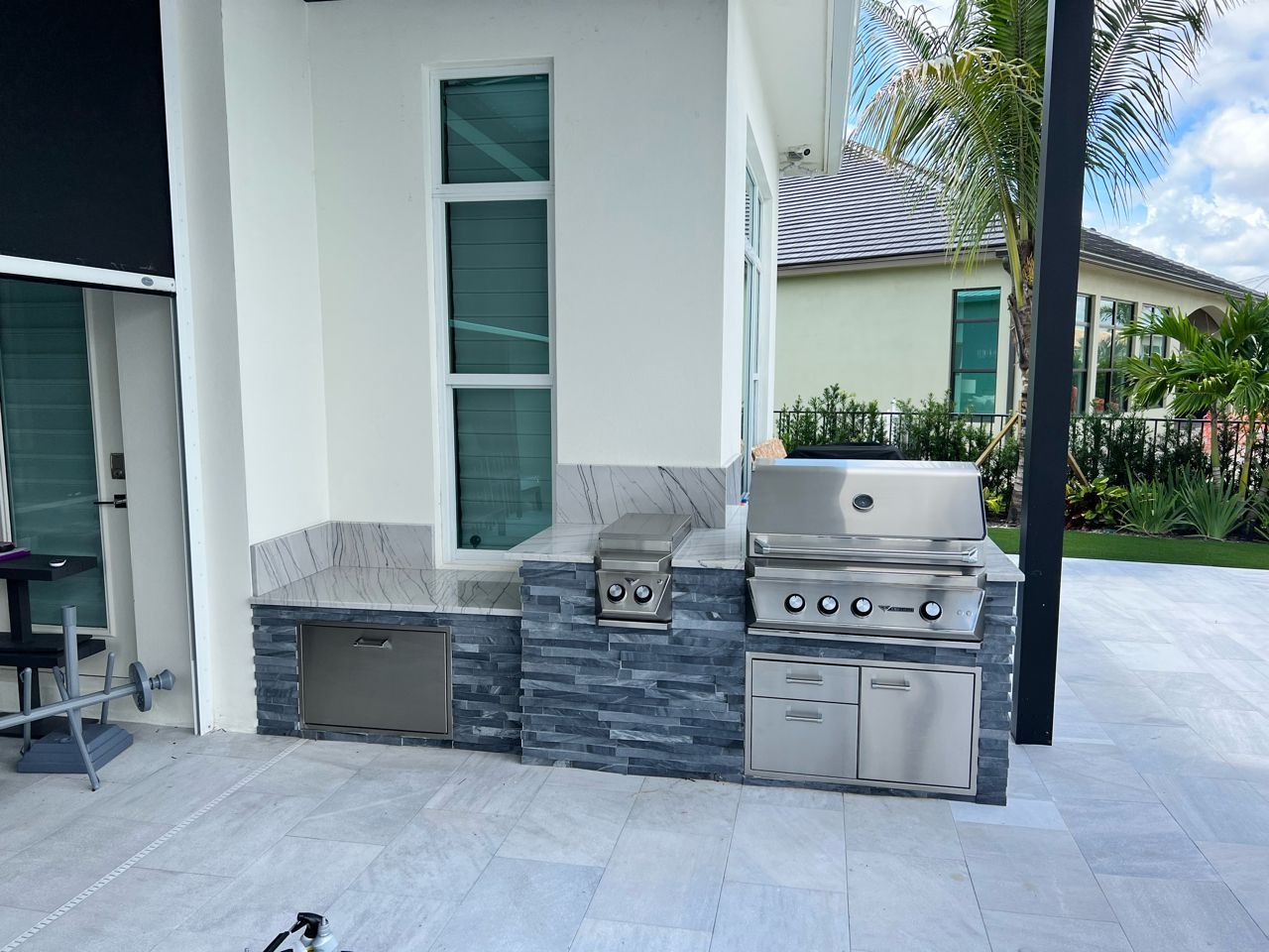 Outdoor built-in grill and sink on a patio beside a white house with gray stone base.