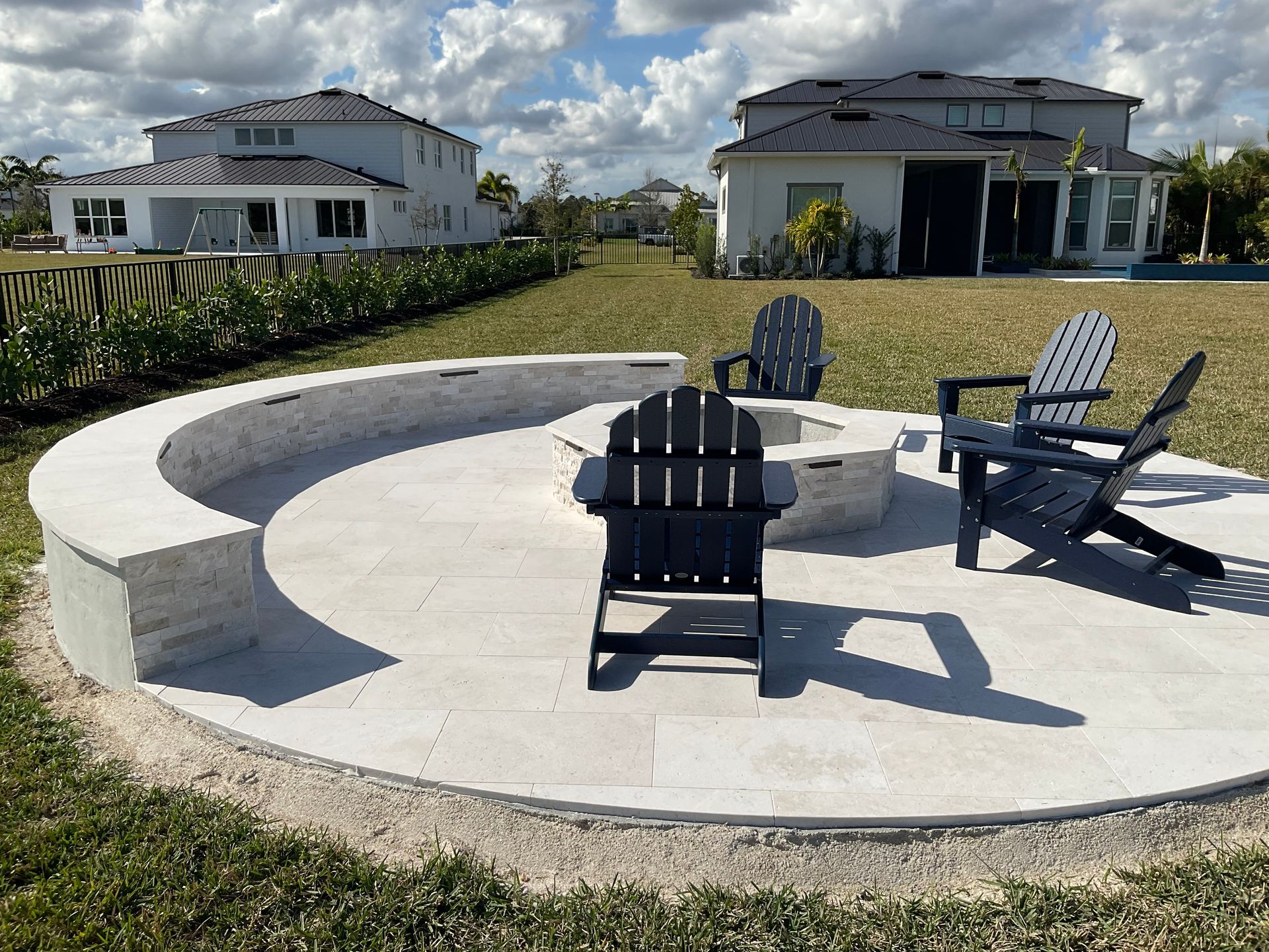 Backyard fire pit area with Adirondack chairs on a concrete patio, lawn, and houses in the background