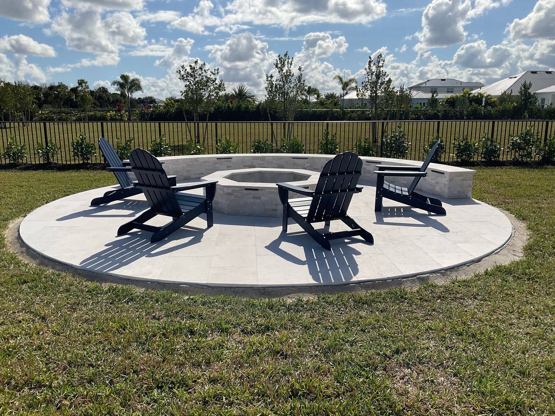 Round white fire pit with black lounge chairs on a grassy patio under a sunny sky.