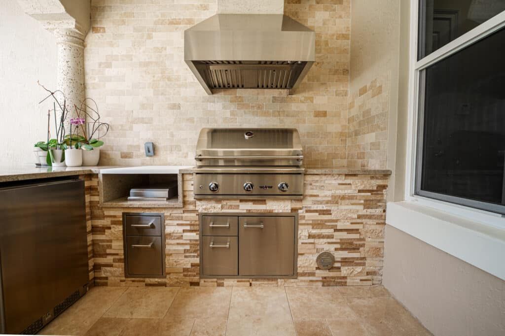 Modern outdoor kitchen with stainless steel grill, hood, sink, and beige stone tile walls