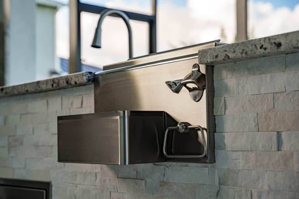Wall-mounted stainless steel paper towel dispenser and faucet in a tiled public sink area.