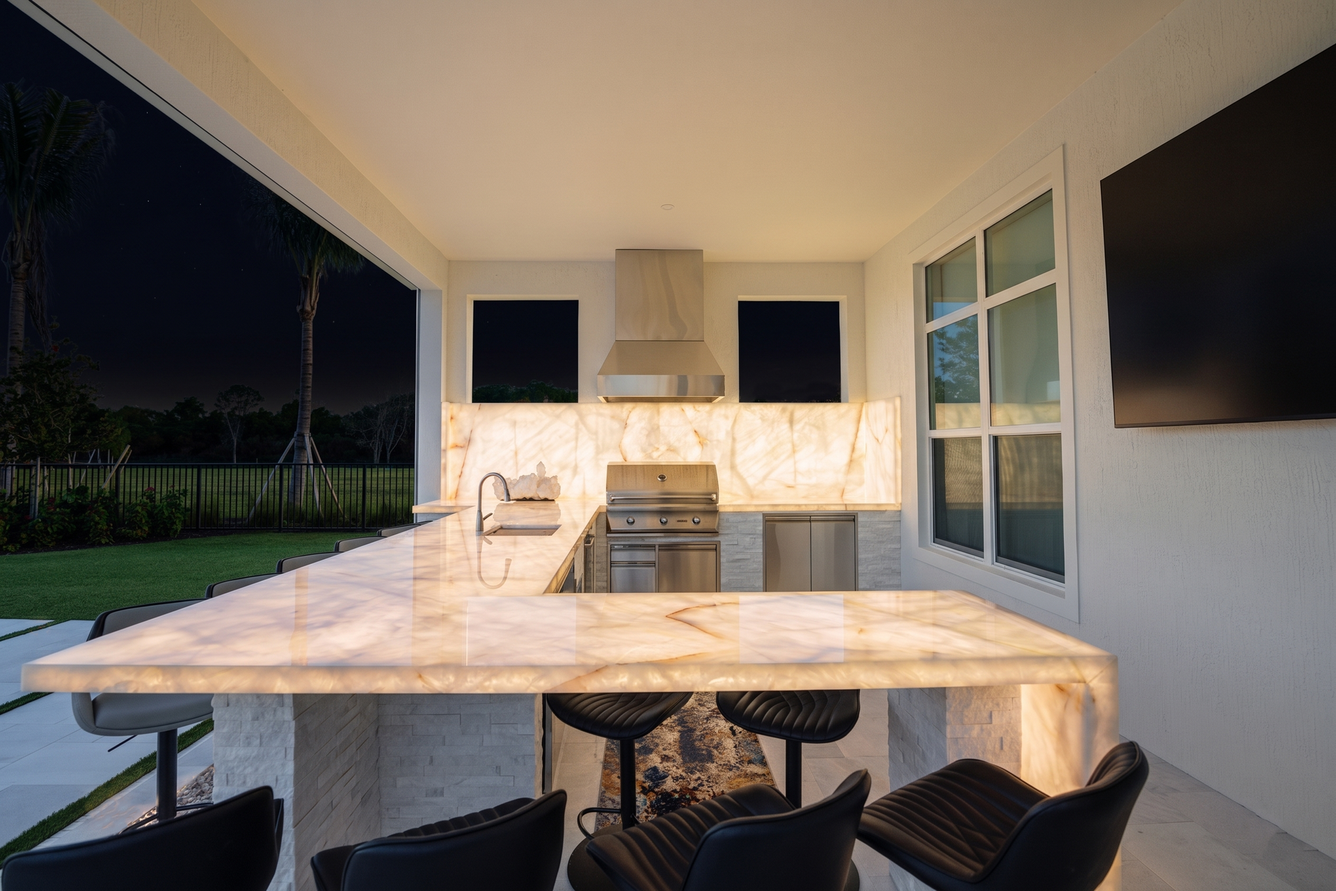 Modern outdoor patio with dining table, built-in grill, and large TV at dusk