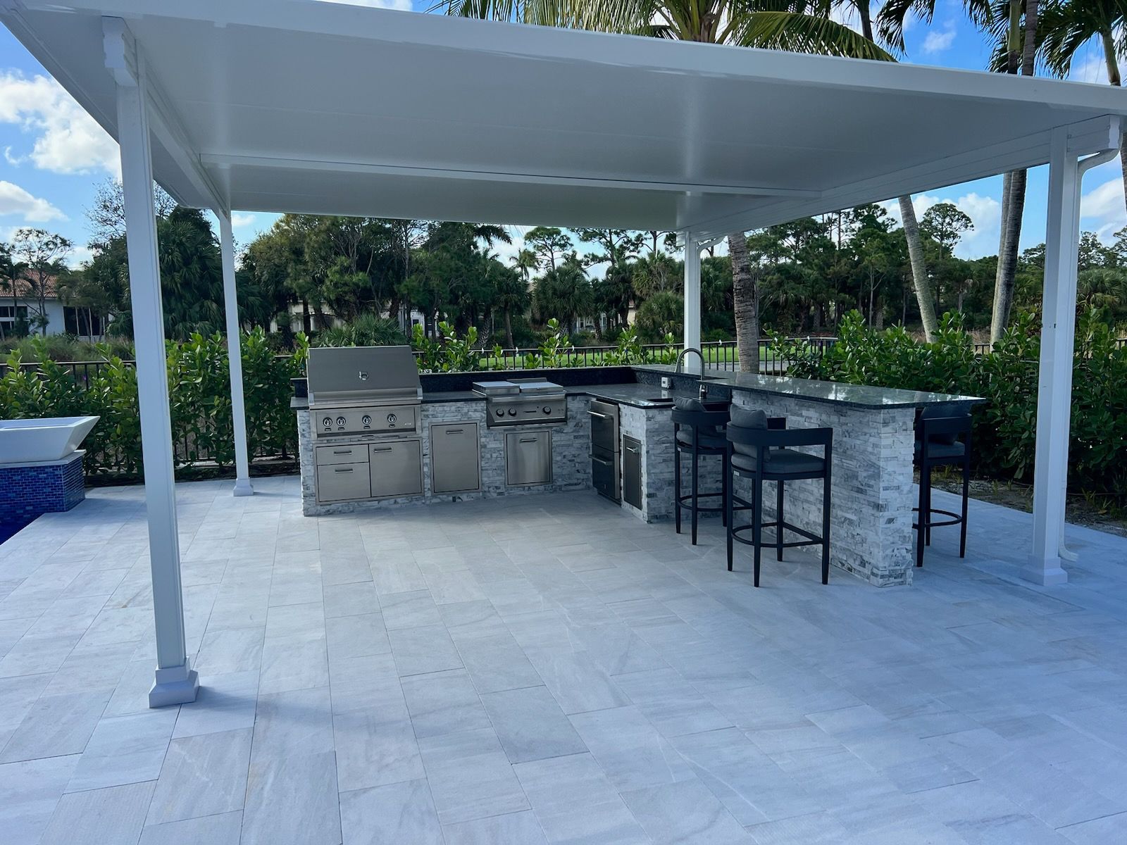 Outdoor patio with white pergola, stone bar, and black bar stools beside a poolside garden