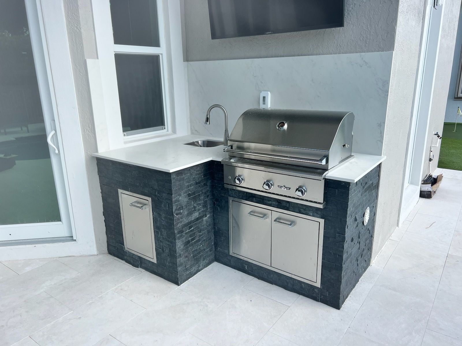 Built-in outdoor grill and sink on a patio with dark tile and light stone counters