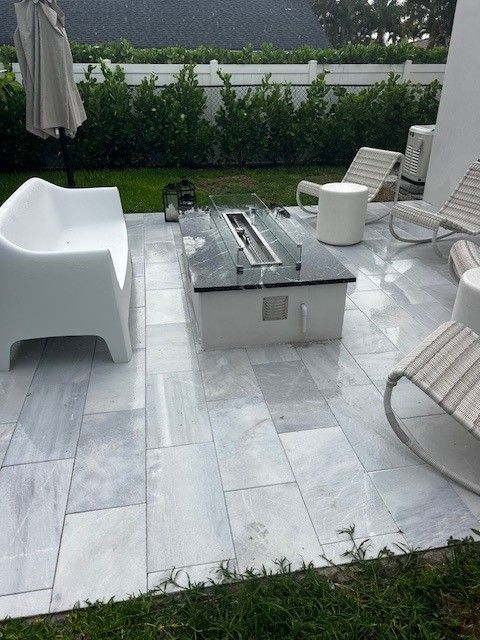 Modern patio with white outdoor seating, gray stone tiles, and a central fire pit near a hedge.