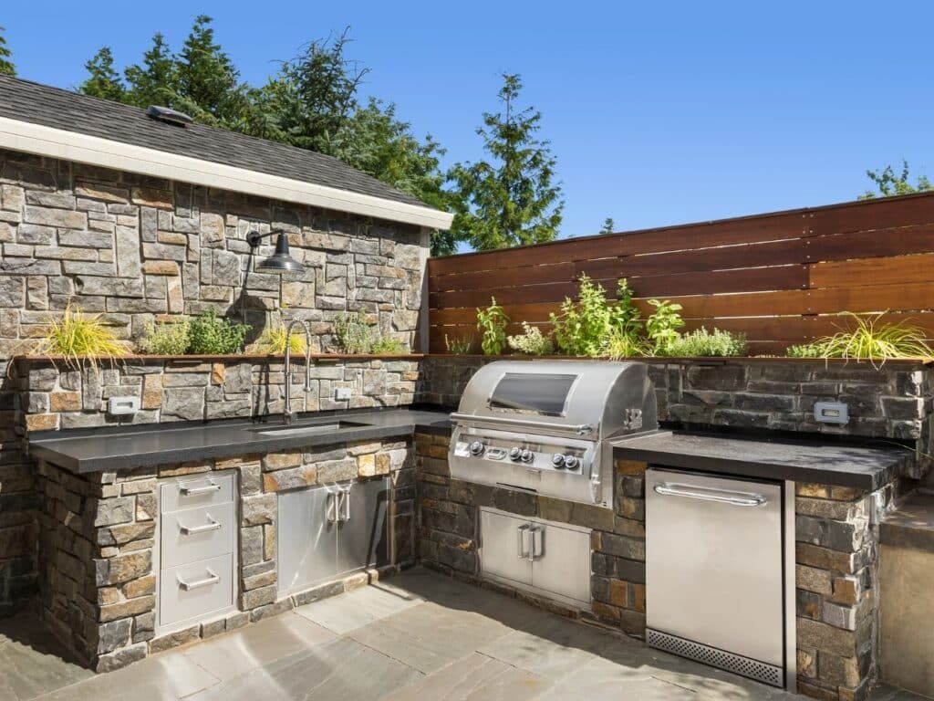 Outdoor stone backyard kitchen with grill, sink, and cabinets under a blue sky