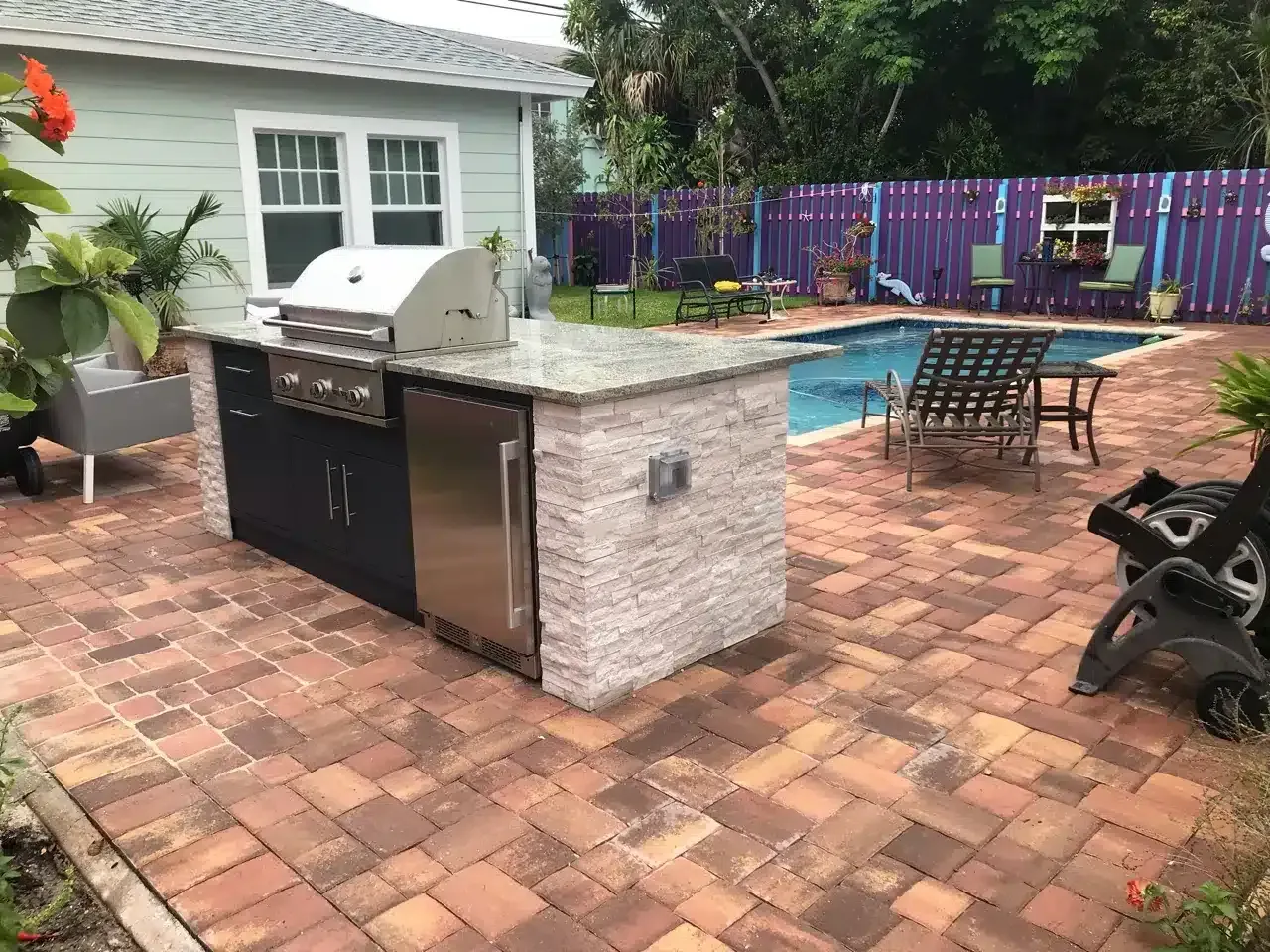 Backyard patio with brick pavers, grill island, pool, chairs, and colorful fence.