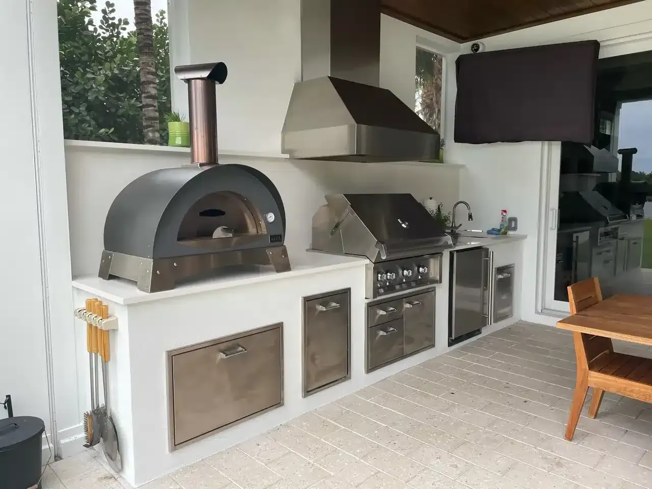 Outdoor patio kitchen with built-in pizza oven, grill, sink, and TV under a covered roof